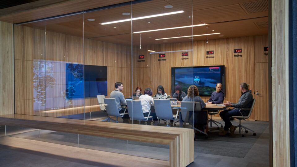 Eight people gathered around a long table in a conference room with two large screens on two walls and digital clocks above one of the screens on the wall. The meeting is visible through a wall of glass.