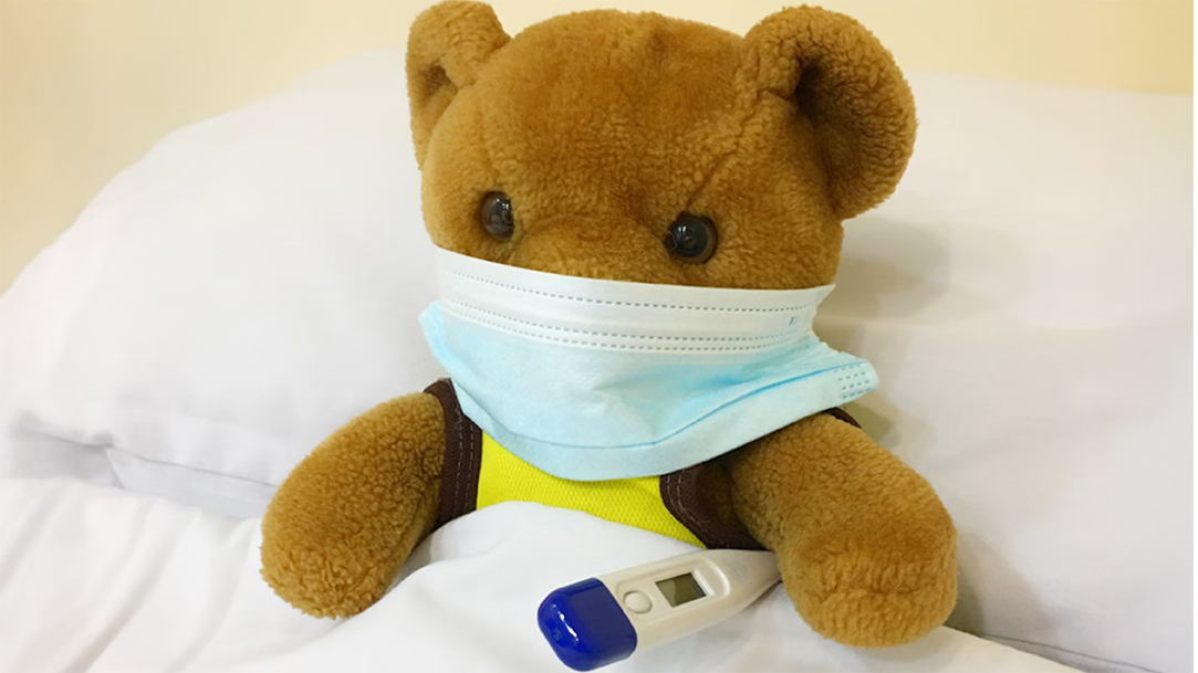 An image of a teddy bear with cold symptoms, tucked in a bed wearing a mask and with a thermometer