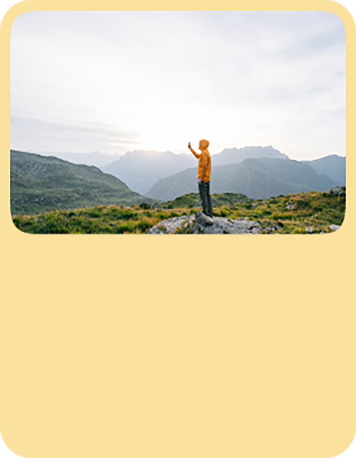 Person in a yellow jacket standing on a rocky hilltop, holding a phone against a backdrop of green valleys and distant mountain peaks under a bright sky