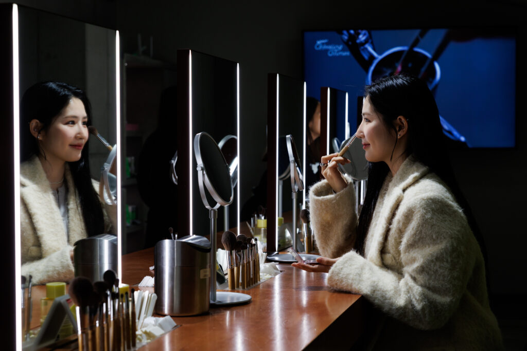 A woman with long black hair and wearing a white jacket looks at herself in the mirror as she uses a make-up brush on her face.
