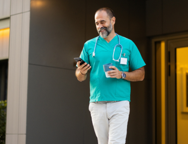Healthcare worker in scrubs walking outside a building, checking a phone.