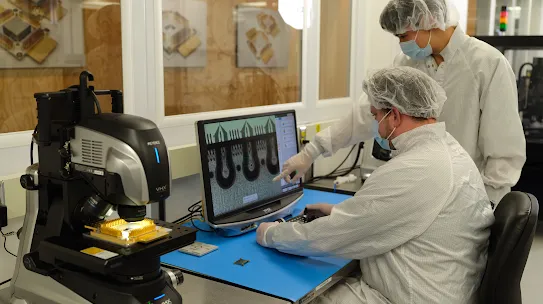 Two scientists in lab coats, hairnets, and face masks examine a computer screen displaying microchip details, with one pointing at the screen.